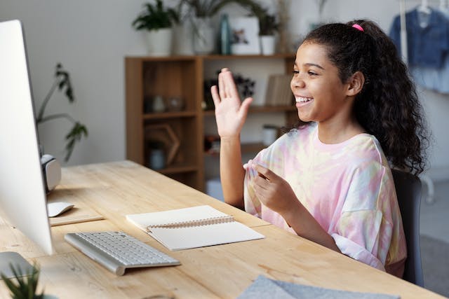 Girl in pink shirt waves at the computer camera.